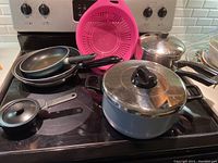 Photo showing all items arranged on stove top including two frying pans, large grey pot with lid, double boiler, pink plastic strainer, and single egg poacher.