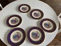 Six decorative plates arranged on table showing blue and gold rims and Victorian illustrations in center.