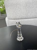 Clear faceted crystal cat figurine with black eyes, silver metal tail and whiskers on black surface with a white chair background.