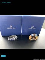 Two Swarovski crystal paperweights displayed side by side in front of their original blue boxes with Swarovski logo.