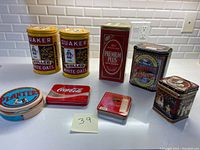 View of all seven assorted tins including 2 Quaker Oats tins, Premium Plus, Bassett's, tea, Planters nut, and Coca-Cola tins arranged on counter.