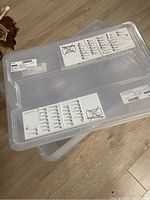 Three clear plastic IKEA Samla storage bins stacked on a wooden floor, showing lids with warning and instruction stickers.