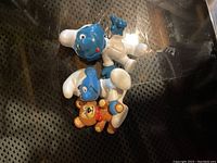Photo showing three Smurf figurines; two Baby Smurfs in white hats and pants with blue skin, one near a small orange teddy bear toy.