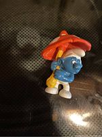 Photo of a small plastic Grumpy Smurf figurine holding a red and white spotted mushroom umbrella.