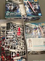 Two HG 1/144 Bandai Gundam model kit boxes, one open showing sprues with plastic parts in white, red, black, and blue, plus instruction sheets. Box edges show damage.