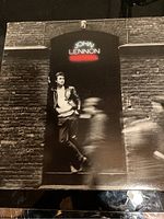 Front cover of John Lennon 'Rock 'n' Roll' vinyl LP showing black and white image of John Lennon leaning on brick wall with album title in neon style text.