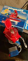 Red Playmobil vintage scuba submarine toy with visible mechanical arms, cockpit, and attached propulsion device placed in front of original box featuring underwater scenery.