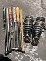 Five baseball bats of various brands and materials, aligned side by side, accompanied by two black Allegro umpire shin guards. Floor surface is rough concrete.