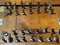 Photo showing all silver-toned Roman/Greek style chess pieces arranged with pawns, knights, bishops, rooks, queen and king visible, placed on a wooden table.