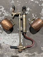 Two copper mugs and a vintage keg tap on a rough concrete floor. The mugs are hammered copper with brass handles. The keg tap is metallic with patina, rubber hose, and black grips.