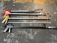 Five reachers laid on a concrete floor, showing different colored grips and metal shafts with claw jaws at the end.