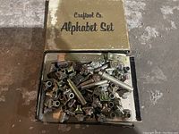 Box with multiple metal press stamps labeled 'Craftool Co. Alphabet Set'. Various metal letter stamps and holders visible inside box.