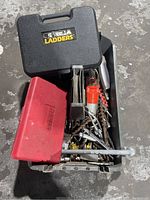Top view of open metal tray filled with assorted drill bits and hardware, with black Gorilla Ladders and red Parker plastic cases on top
