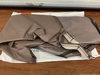 Brown barbecue cover folded on a wooden surface showing adjustable strap and plastic buckle.