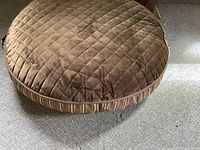 Large round brown dog bed with quilted pattern on top and striped sides on carpeted floor.