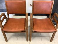 Front view of two mid-century modern occasional chairs side by side showing wooden frames and brown leather upholstery.