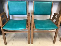 Two turquoise vinyl upholstered mid-century modern style chairs with wooden armrests and frames, showing overall design and colors.