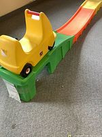 Yellow ride-on cart positioned on green base ramp section of roller coaster track on carpeted floor.