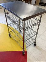 Front diagonal view of the rolling kitchen cart showing stainless steel top and wire shelves with casters on floor.