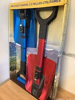 Pair of blue and red utility shovels in original packaging standing upright, showing foldable handles and blade colors.