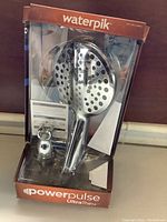 Front view of Waterpik PowerPulse UltraThin+ shower head in original packaging showing shower head, hose, and mounting bracket inside brown and clear box.