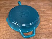 View of the skillet underside showing Tramontina branding and size marking, glossy turquoise enamel finish on cast iron body.