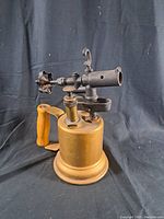 Photo showing the vintage bronze torch tool from a front angle, highlighting the cylindrical base, wooden handle, and metal nozzle with adjustment valve.