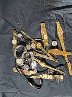 Lot of multiple wristwatches spread on black fabric background showing variety of watch styles and bands, mostly metal, some gold-tone and silver-tone.