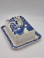Butter dish with lid on showing ornate blue and white china design featuring trees and building pattern. Rectangular shape with handle on top of lid.