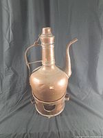 Front and side views of large vintage bronze liquid heater showing patina and wear. The spout, handle, and base stand are visible.