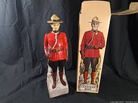 Front view of the vintage Canadian Mountie statue standing next to its original box labeled 'Canadian Mist'. The Mountie is dressed in red serge uniform with a brown hat and boots.