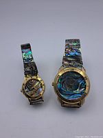 Two vintage brass watches with multicolored shell inlay bands and faces shown side by side.