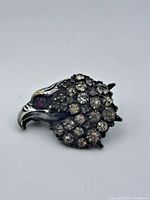 Close-up of front of silver eagle pin. Shows clear rhinestones and a bright red ruby-colored eye stone set in a silver-toned metal shaped like an eagle's head.