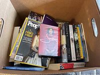 Box containing assorted hard and soft-covered books and DVDs. Visible items include commemorative People magazine featuring Prince Philip, 'Suffragette' DVD, 'The Crown: Official Companion', various fiction and non-fiction books.