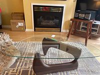 Front view of coffee table showing triangular wood veneer base supporting a clear glass top on a patterned rug