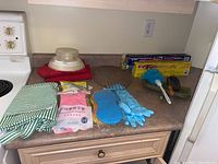 Countertop showing green and white striped apron, pack of new pink latex gloves, rolls of tin foil and parchment paper, blue duster, green dustpan and brush, IKEA kitchen scale, and oven gloves.