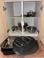Kitchen cabinet containing a stainless steel mixing bowl, two pots with lids, a small fry pan, green non-stick fry pan, and a roasting pan with rack placed below.
