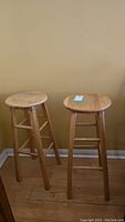 Two light natural wood stools, round top, four legs with support bars, positioned against a plain yellow wall on hardwood floor.