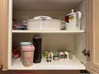 Two shelves inside a kitchen cabinet showing the Christmas-themed teapot, CorningWare casserole dish and bowls on the top shelf, and egg cups, salt and pepper shakers, and travel mugs on the bottom shelf.