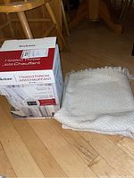 Photo of Brookstone Heated Throw in original box next to cream woven throw on hardwood floor.