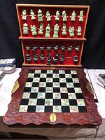 Full view of the Asian chess set showing the green and black carved chess pieces in a red display case above the chessboard with carved wooden frame and brass handles.