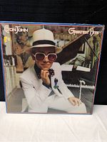 Front cover showing Elton John in a white suit and pink glasses sitting in front of a piano