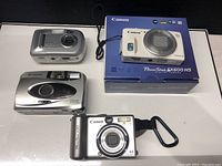 Overview of all four cameras: Canon PowerShot SX600 HS (white, on box), Canon PowerShot A80 (silver), Polaroid 470 AF (silver), Kodak camera (silver).