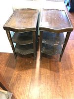 Two antique wooden side tables shown from above and front angle on a hardwood floor. Both have curved front edges, raised sides, and two lower curved shelves with worn paint and scratches.