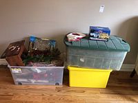 Wide shot of three plastic totes, a wooden stool, and assorted Christmas decorations arranged on a hardwood floor against a beige wall.