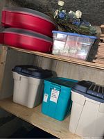 Three large storage totes and wreath containers stacked on shelves with artificial floral decorations on top