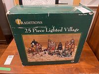 Box front showing 25 piece lighted village with buildings, figures, and accessories in green box with Traditions logo.