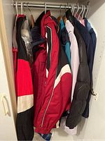 View of 8 winter coats hanging closely on a rack in a closet, colors include red, black, blue, grey, and white, styles appear winter insulated and puffer