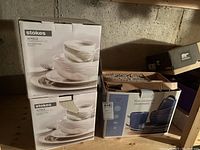 Two boxes of Stokes 16-piece stoneware dinnerware sets in white/gray marble pattern stacked on wooden shelf with a box of Sonoma blue stoneware dinnerware set nearby