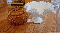 Two bowls on wood floor: white milk glass pedestal bowl with ruffled edge and amber glass lidded bowl with ribbed pattern.